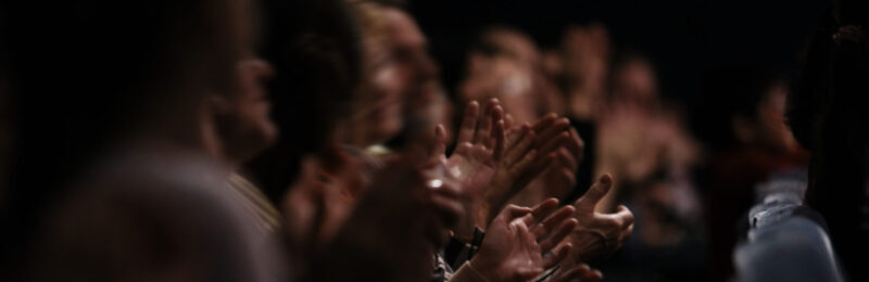 View down a row of people sitting in an audience of people clapping their hands in appreciation of a performance